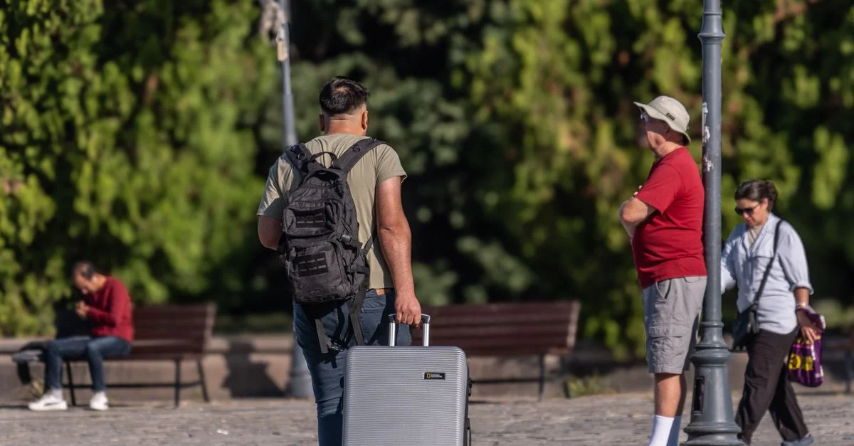 Un homme avec une valise et un sac à dos se promenant dans un parc ensoleillé à İzmir, Turquie.