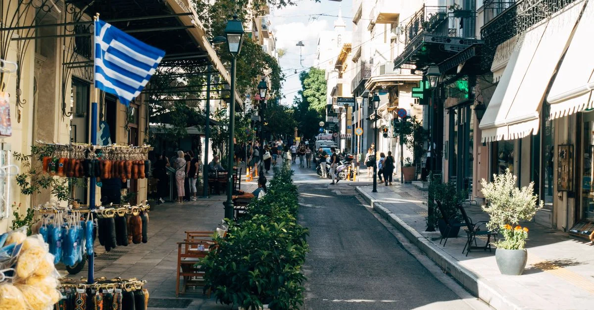Rue ensoleillée en Grèce bordée de boutiques et d'un drapeau grec, capturant la vie urbaine vibrante.