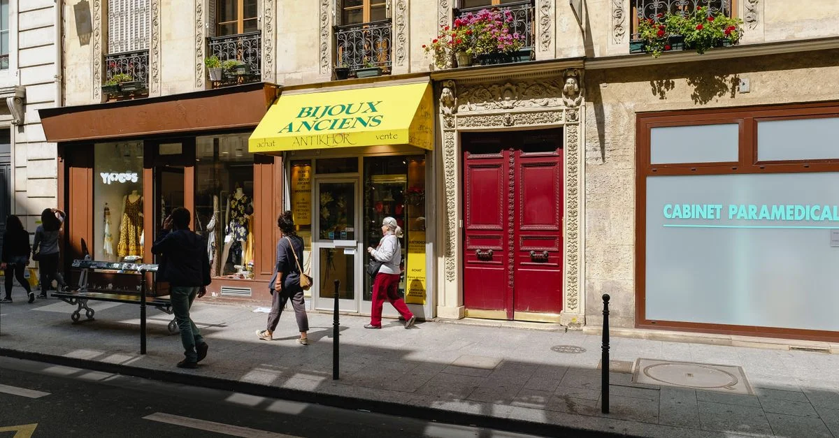 Des personnes passant devant de charmantes boutiques parisiennes par une journée ensoleillée, mettant en valeur l'architecture vintage et la vie animée de la rue.
