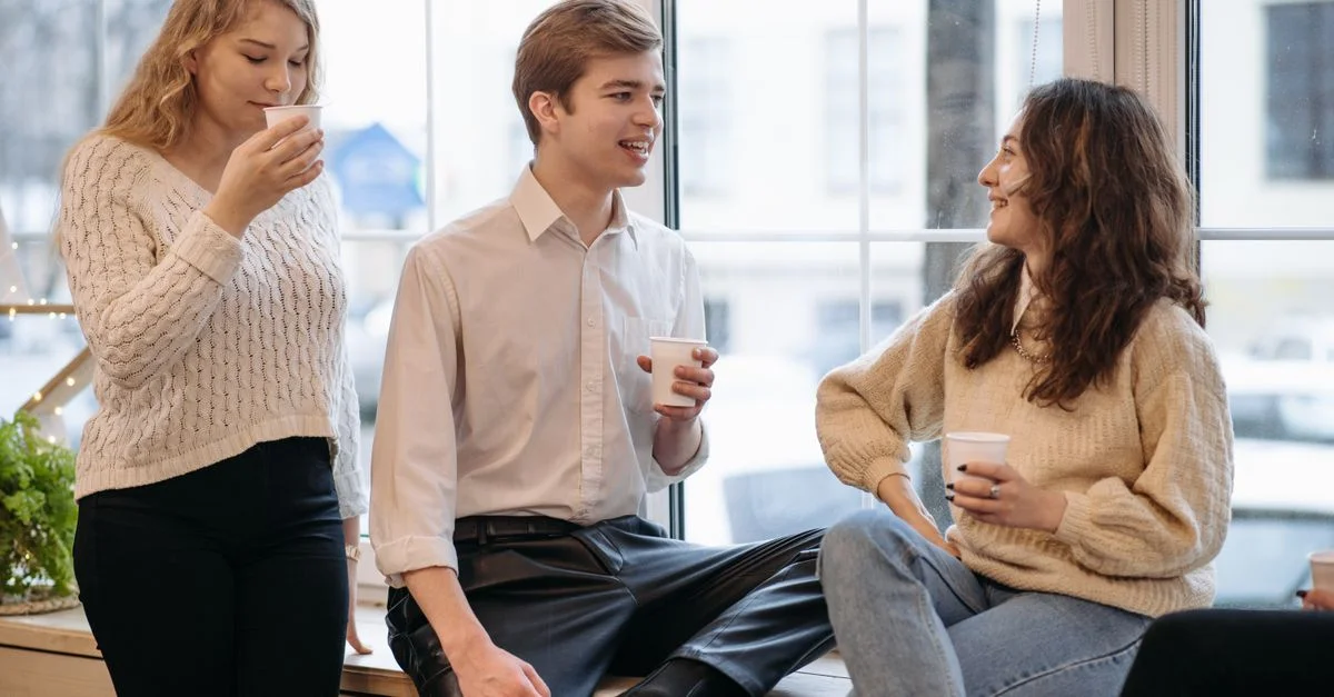 Trois jeunes adultes profitant d'un café et d'une conversation dans un cadre intérieur détendu près de la fenêtre.