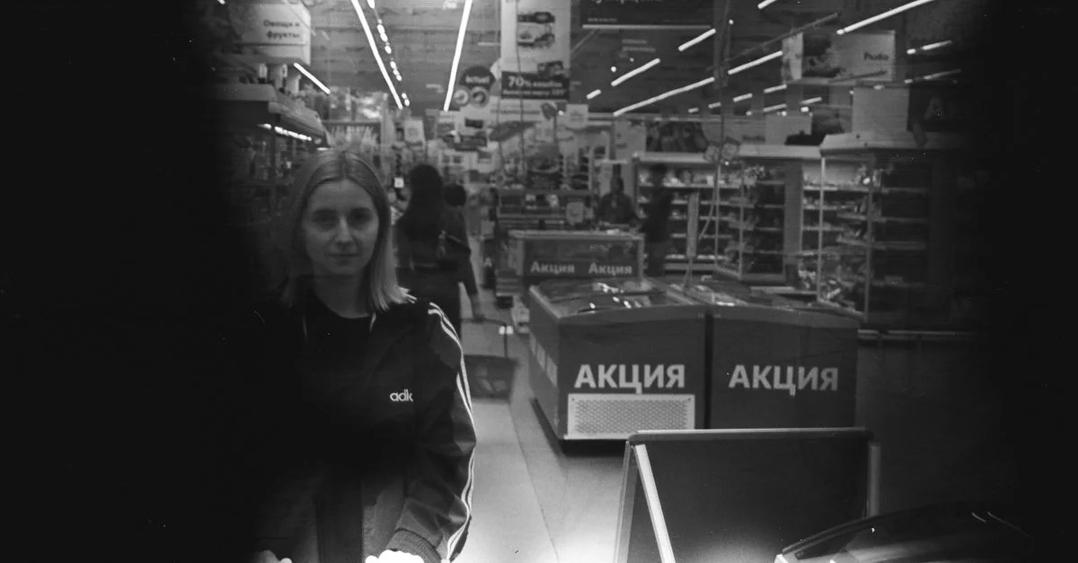 Une femme faisant ses courses dans une allée de supermarché en niveaux de gris, poussant un chariot.