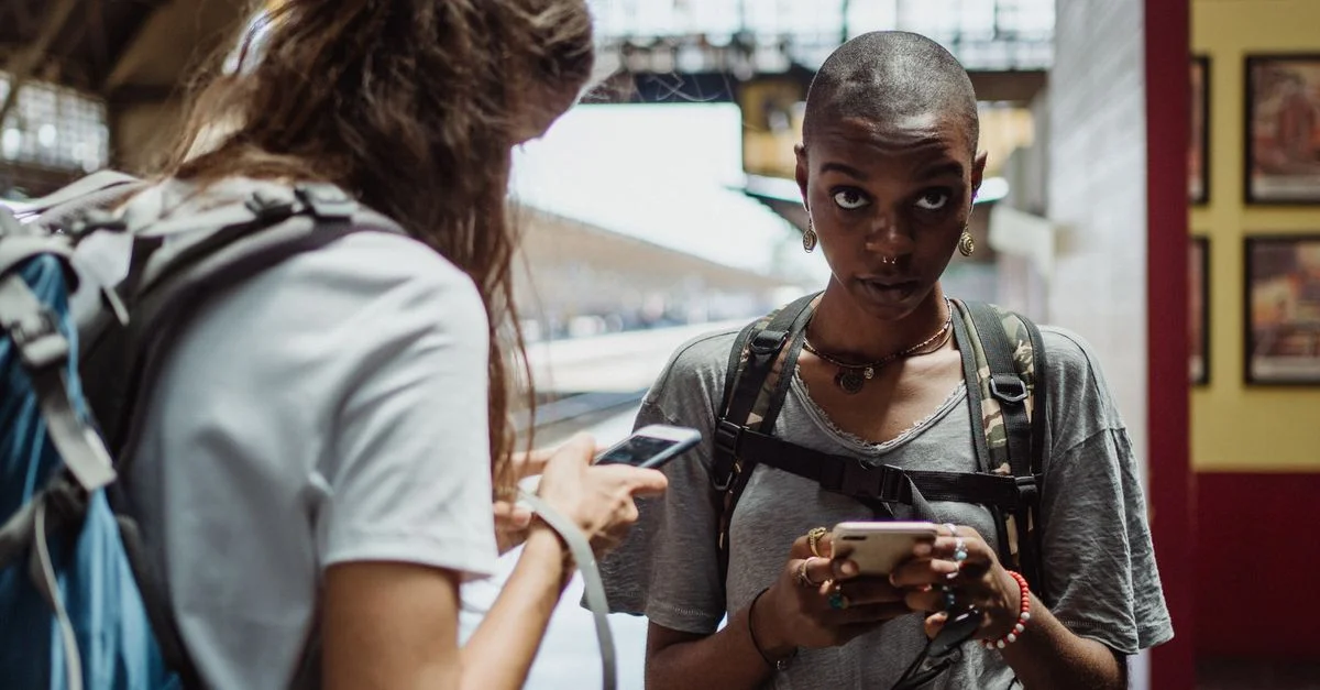 Deux voyageuses diversifiées utilisant des smartphones dans une gare, sacs à dos visibles.