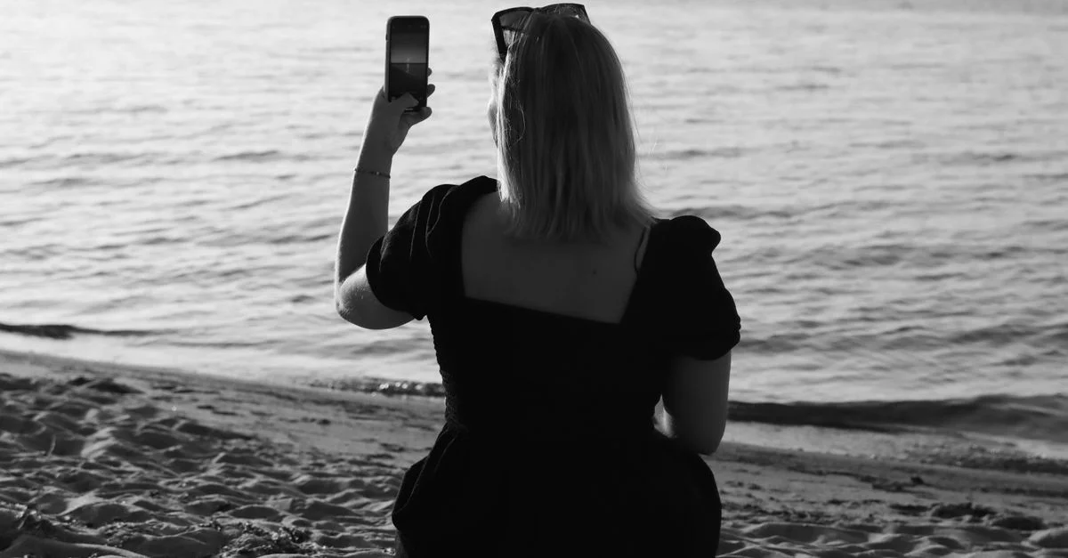 Une femme portant une robe noire utilise un smartphone pour capturer le paysage de plage serein à Jönköping, en Suède.